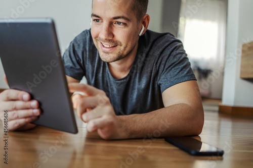 Online tablet research. A happy male person lies on the floor in the room and checks the news in the world. He uses a tablet and scrolls the touch screen with his finger. Next to it is a telephone