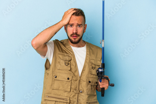 Young caucasian fisherman with tattoos holding rod isolated on blue background being shocked, she has remembered important meeting.