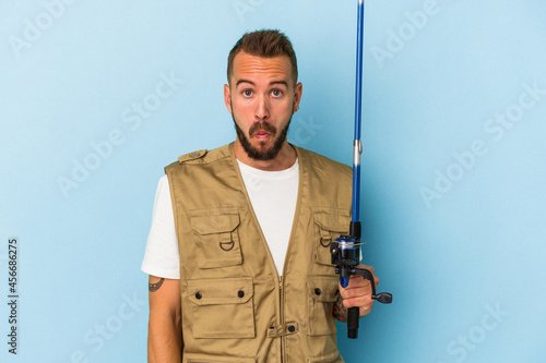 Young caucasian fisherman with tattoos holding rod isolated on blue background shrugs shoulders and open eyes confused.
