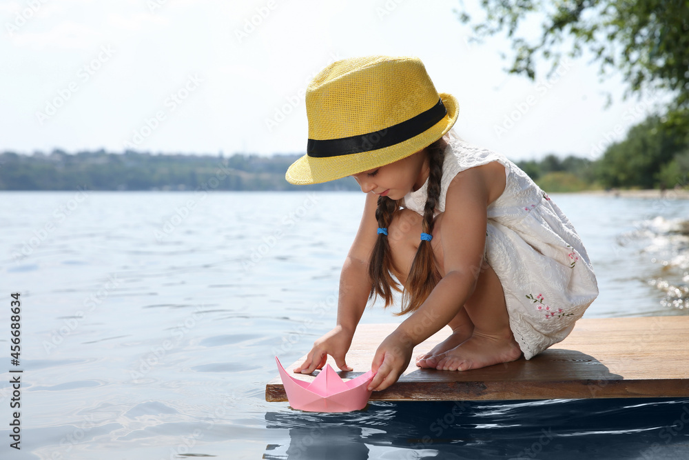 Cute little girl playing with paper boat on wooden pier near river Cute little girl playing with paper boat on wooden pier near river