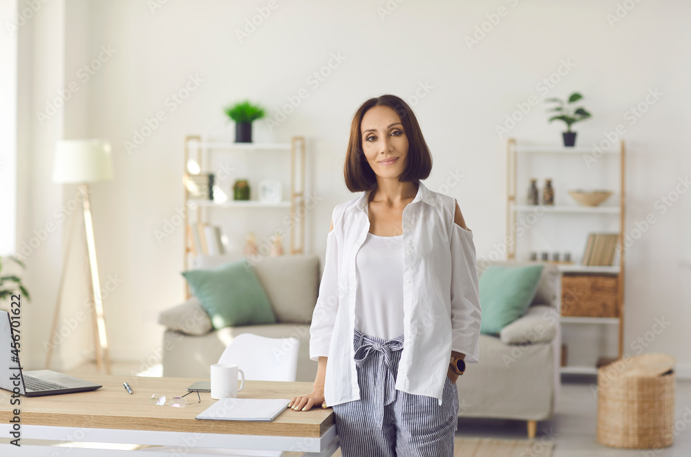 Portrait of confident middle-aged business woman standing in her cozy home office. Beautiful and stylish Caucasian woman in casual clothes stands at her desk and smiles looking at camera. Portrait of confident middle-aged business woman standing in her cozy home office. Beautiful and stylish Caucasian woman in casual clothes stands at her desk and smiles looking at camera.