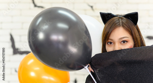 Portrait of young beautiful asian woman wearing witch costume posing with balloons on white brick background decorated with black paper bat shape in halloween party - Free space for copy text