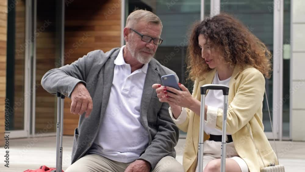 Beautiful mature couple with suitcases is sitting on a bench waiting for their flight. A man and a woman are discussing what they saw on the screen of a mobile phone. Beautiful mature couple with suitcases is sitting on a bench waiting for their flight. A man and a woman are discussing what they saw on the screen of a mobile phone.