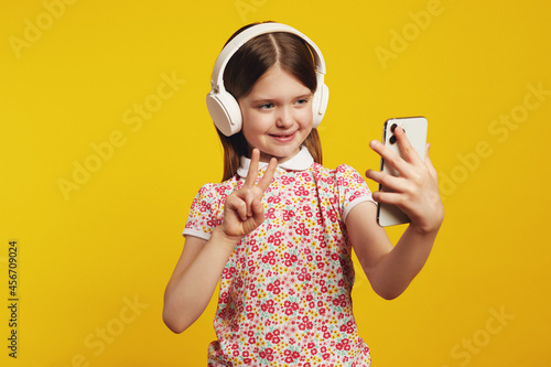 Caucasian girl shows peace gesture, uses smartphone, listens favorite music via white headphones wears summer clothes, isolated over yellow background photo