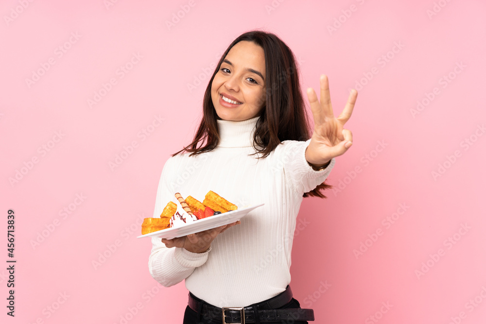 Young brunette woman holding waffles over isolated pink background happy and counting three with fingers Young brunette woman holding waffles over isolated pink background happy and counting three with fingers