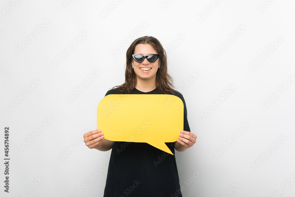 Young cheerful woman wearing sunglasses is holding a yellow speech bubble over white background. Young cheerful woman wearing sunglasses is holding a yellow speech bubble over white background.