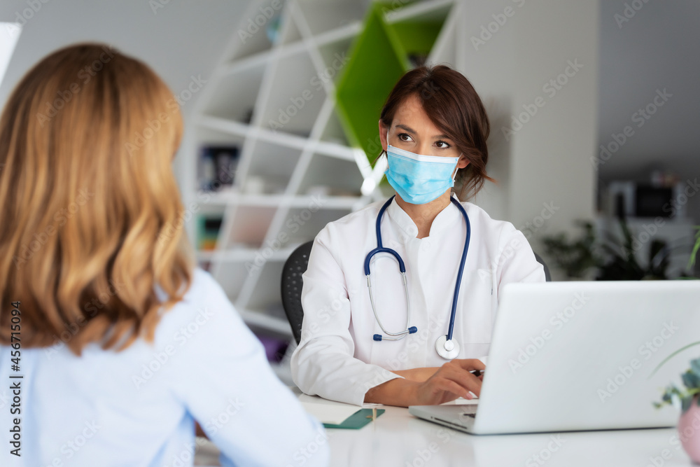 Female doctor wearing face mask and consulting her patient while sitting at desk in doctor's office Female doctor wearing face mask and consulting her patient while sitting at desk in doctor's office