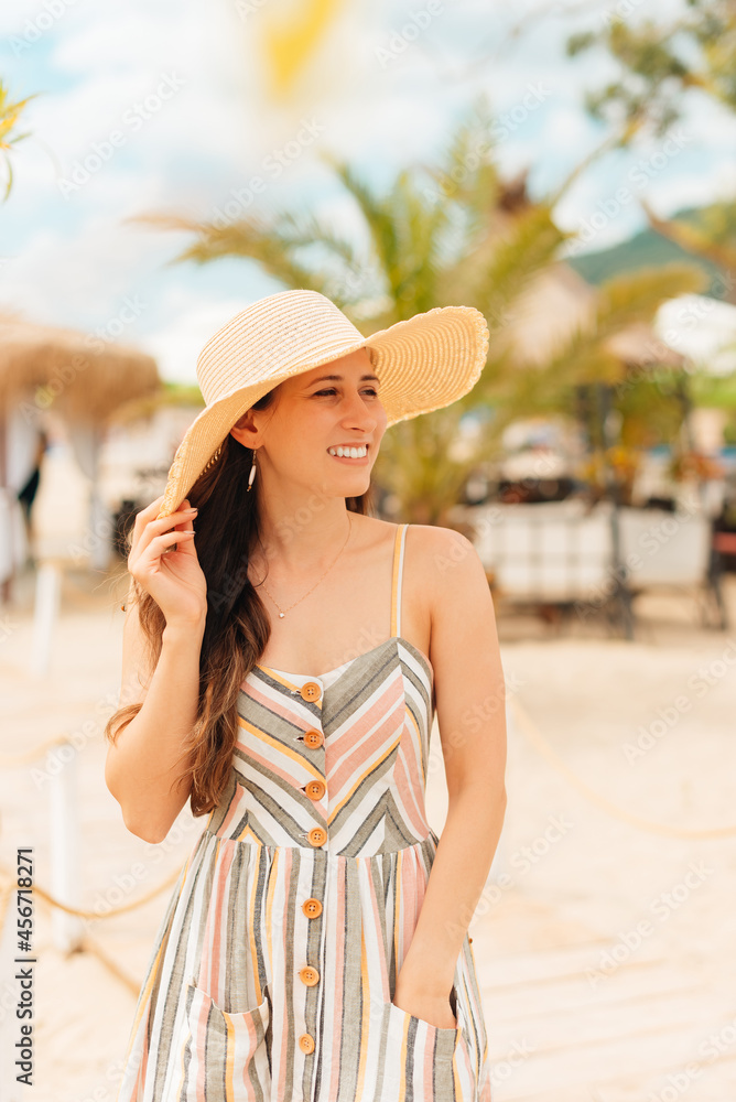 Vertical photo of a young woman on vacation on the beach, wearing a hat and dress.