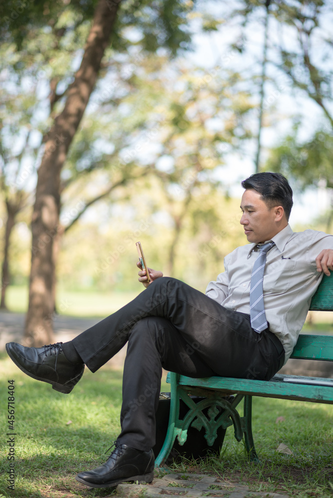 Businessman sitting on bench and using mobile phone in park Businessman sitting on bench and using mobile phone in park