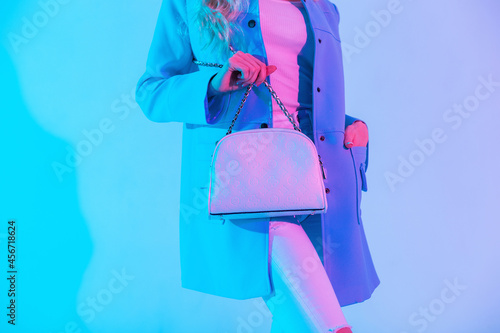 Beautiful stylish woman in colored blue blazer, blouse and jeans with white fashionable handbag in the studio at neon pink light. Close-up of a leather female bag