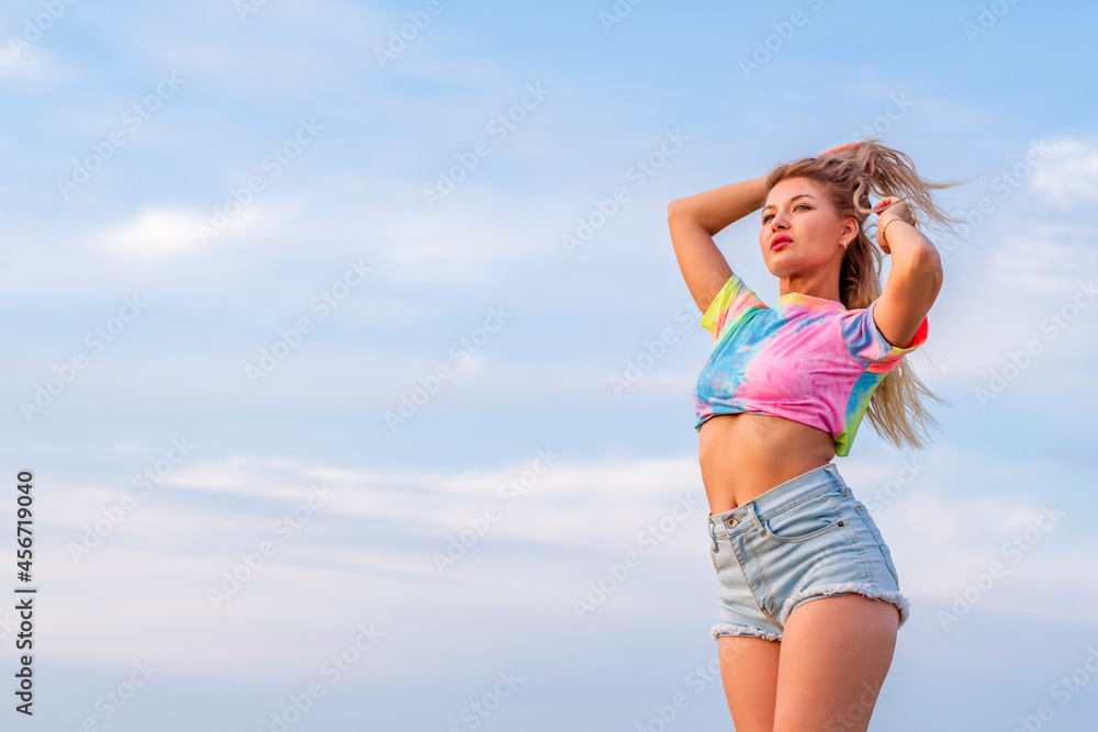 Young girl in bright casual clothes stands half-turned to the camera against the background of a blue evening summer sky. Looks into the distance.