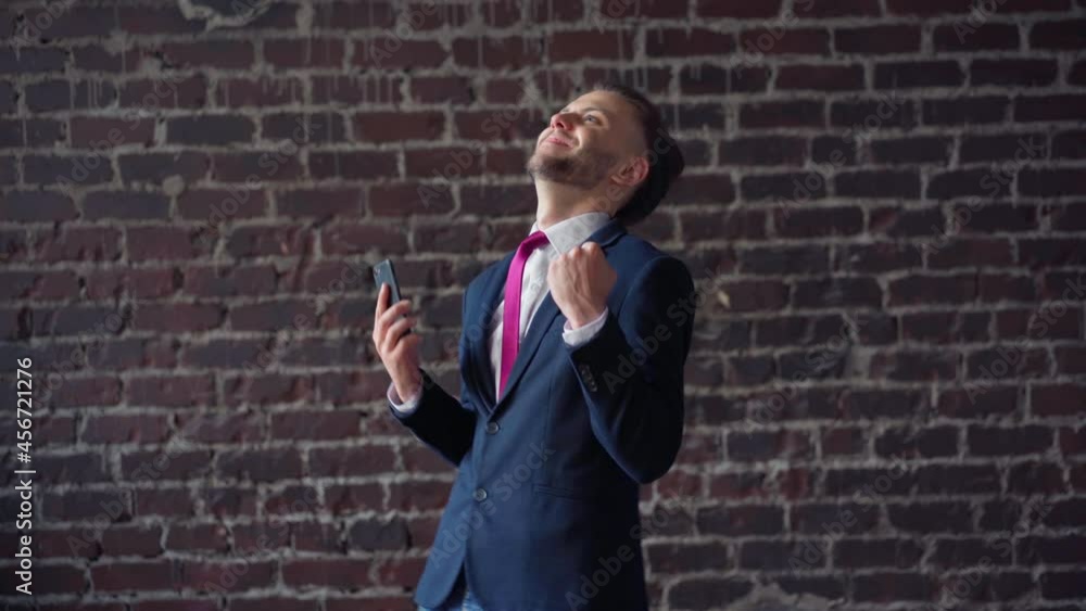Surprised businessman reading good news on phone indoor red brick wall background. Middle shoot dancing happy male professional enjoying success. Smiling business man dance dressed business suit Surprised businessman reading good news on phone indoor red brick wall background. Middle shoot dancing happy male professional enjoying success. Smiling business man dance dressed business suit