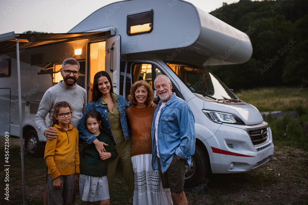 Portrait of multi-generation family looking at camera and smiling outdoors at dusk, caravan holiday trip. - obrazy, fototapety, plakaty 