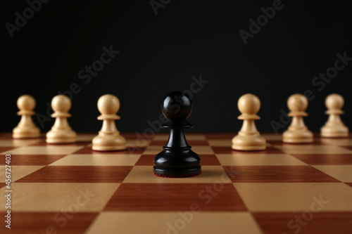 Black pawn in front of white ones on wooden chess board against dark background