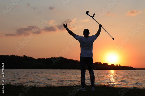Man raising elbow crutch up to sky near river at sunset, back view. Healing miracle