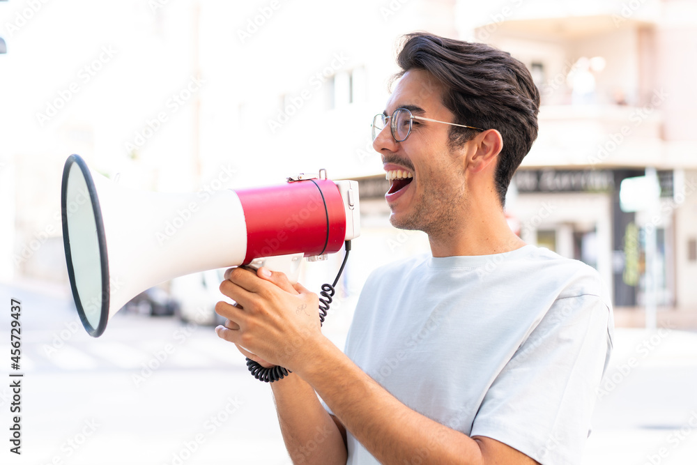 Young caucasian man at outdoors shouting through a megaphone to announce something in lateral position Young caucasian man at outdoors shouting through a megaphone to announce something in lateral position