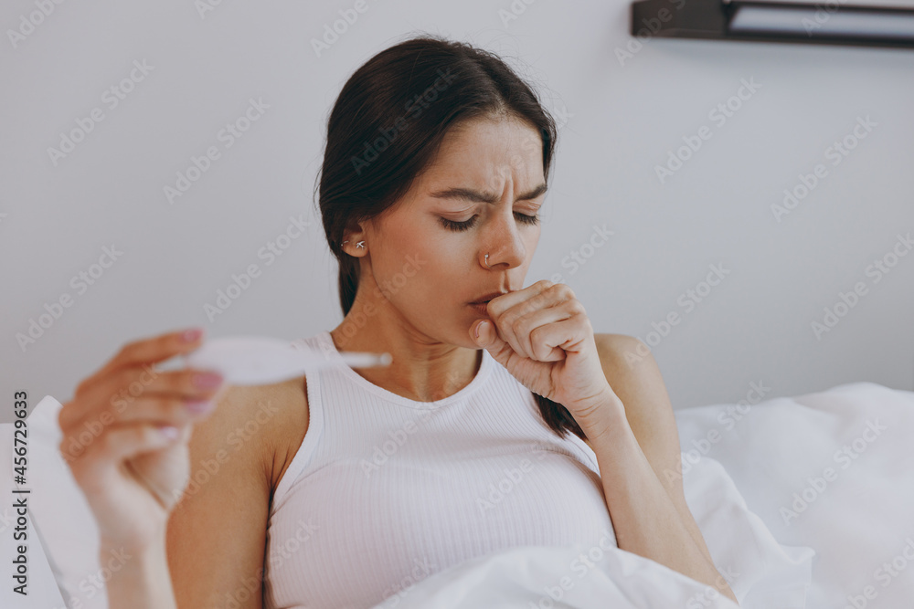 Sick ill depleted young woman in white tank top sit in bed hold in hand thermometer coughing in bedroom lounge home in own room house wake up. Healthy lifestyle disease treatment cold season concept. Sick ill depleted young woman in white tank top sit in bed hold in hand thermometer coughing in bedroom lounge home in own room house wake up. Healthy lifestyle disease treatment cold season concept.