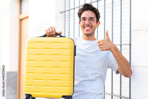 Young caucasian man at outdoors in vacation with travel suitcase and with thumb up