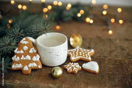 Christmas gingerbread cookies, coffee cup, fir branches and bauble on background of warm lights bokeh on rustic wooden table. Moody atmospheric image. Winter countryside hygge. Seasons greeting