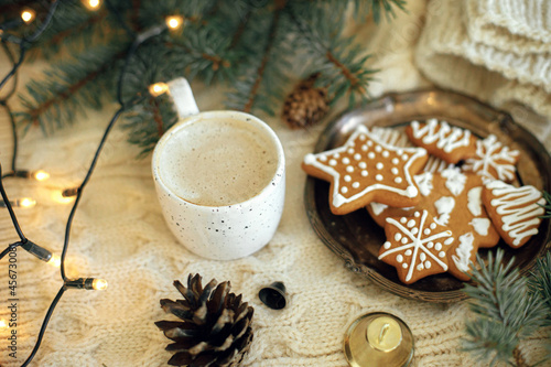 Homemade christmas gingerbread cookies, warm coffee in stylish cup, fir branches and warm lights on cozy knitted background. Atmospheric festive time and hygge home. Happy Holidays. Hello winter