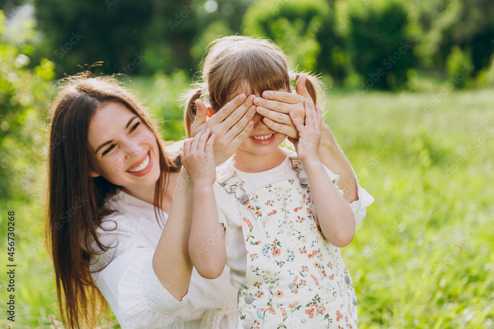 Obraz premium Smiling young woman in white clothes close eyes with hands play guess who or hide and seek with child baby girl 5-6 years old Mommy rest with little kid daughter outdoor together. Love family concept