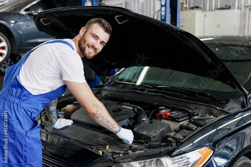 Young professional technician car mechanic man 20s in denim blue overalls white t-shirt gloves fixing problem with raised hood work in modern vehicle repair shop workshop indoor Tattoo translate fun.