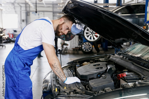 Tired troubleshooter young male professional technician car mechanic man in denim blue overalls white t-shirt fixing problem with raised hood work in light modern vehicle repair shop workshop indoor.