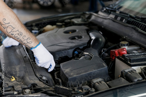 Close up male hands professional technician car mechanic man wearing gloves fixing problem with raised hood look at automobile parts work in vehicle repair shop workshop indoor Tattoo translate fun.