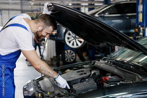 Young professional technician car mechanic man in blue overalls white t-shirt gloves listen music in earphones fix problem with raised hood bonnet work in modern vehicle repair shop workshop indoor.