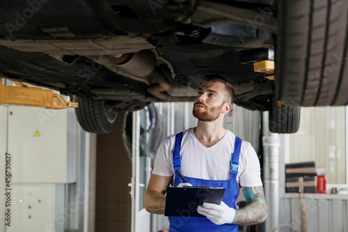 Young serious technician mechanic man wears denim overalls use hold clipboard papers document writing estimate stand near car lift check technical condition work in vehicle repair shop workshop inside