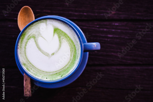 The blue beautiful ceramic cup of hot matcha milk tea which have latte art as a flower with wooden spoon on the surface of the latte on the dark brown wooden table background with coyspace