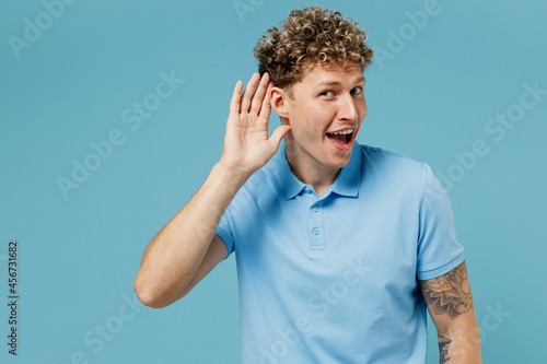 Curious nosy fun vivid energetic young curly man 20s years old wears azure t-shirt try to hear you overhear listening intently hot news isolated on plain pastel light blue background studio portrait.
