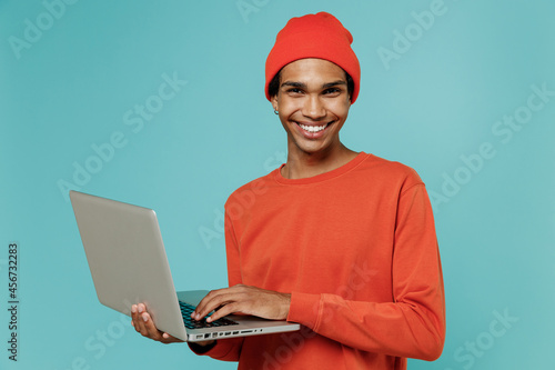 Young smiling happy african american man in orange shirt hat hold use work on laptop pc computer look camera isolated on plain pastel light blue background studio portrait People lifestyle concept