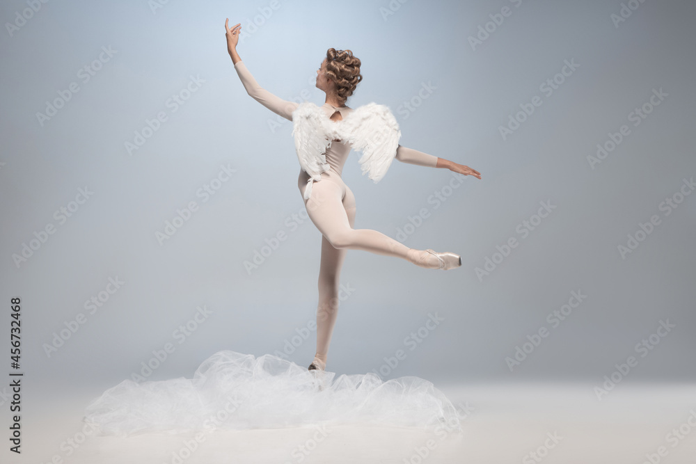 Obraz premium Full-length portrait of beautiful graceful ballerina dancing in image of angel with wings isolated on gray studio background. Art, motion, action, flexibility, inspiration concept.