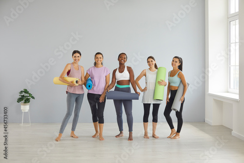 Team of smiling girls in good shape standing in gym and holding fitness mats. Group of happy fit sporty beautiful young multiracial women in leggings and yoga pants posing with mats for sports workout