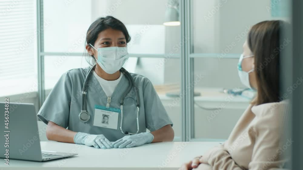 Young Hispanic midwife in medical uniform, protective mask and gloves giving consultation to pregnant woman during prenatal visit in time of coronavirus outbreak Young Hispanic midwife in medical uniform, protective mask and gloves giving consultation to pregnant woman during prenatal visit in time of coronavirus outbreak