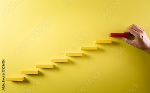Concept of building success foundation. Hand holding wooden block stacking as step stair, Success in business growth concept on yellow background.