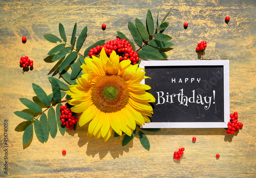 Text Happy Birthday on blackboard. Autumn natural decorations. Yellow sunflower, orange rowan berry with leaves on wooden textured yellow background. Flat lay, top view on wood. Fall greeting card.