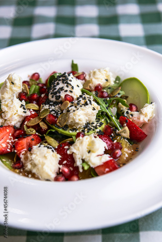 Green salad with cucumber, zucchini and strawberries in a white bowl over green plaid tablecloth.