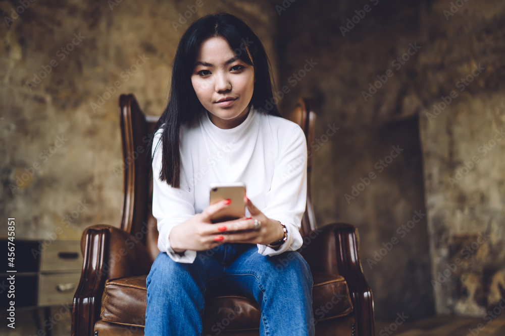 Half length portrait of attractive Asian blogger with modern smartphone gadget in hand looking at camera during leisure pastime for mobile networking, millennial user sitting in chair and posing Half length portrait of attractive Asian blogger with modern smartphone gadget in hand looking at camera during leisure pastime for mobile networking, millennial user sitting in chair and posing