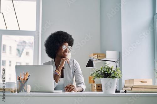 Beautiful young African woman holding hand on chin and smiling while sitting at her working place in office