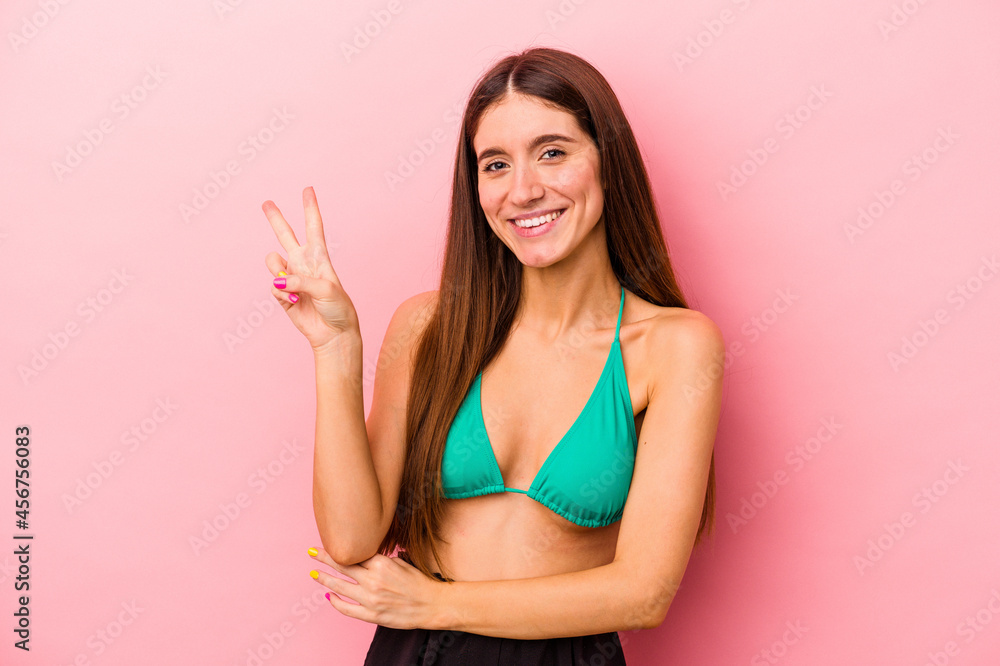 Young caucasian woman wearing bikini isolated on pink background joyful and carefree showing a peace symbol with fingers. Young caucasian woman wearing bikini isolated on pink background joyful and carefree showing a peace symbol with fingers.