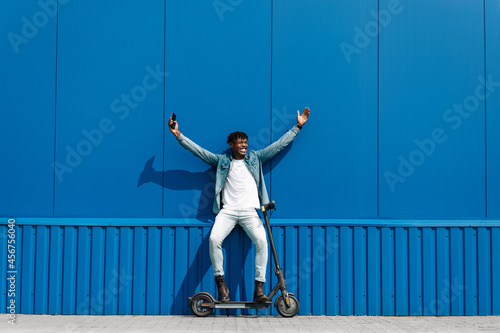black african american man with an electric scooter, shows a gesture of success while holding a phone in his hand
