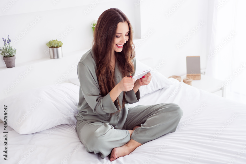 Photo of shiny charming lady grey sleepwear sitting bed legs crossed smiling playing game modern gadget indoors room home