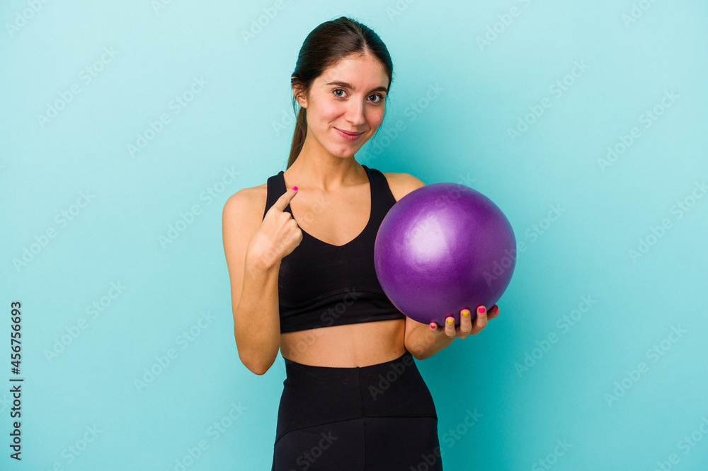 Young caucasian fitness woman holding a ball isolated on blue background pointing with finger at you as if inviting come closer. Young caucasian fitness woman holding a ball isolated on blue background pointing with finger at you as if inviting come closer.