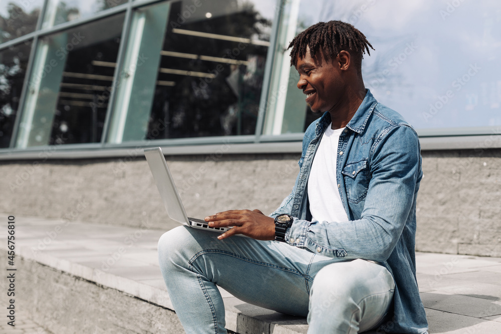 black african american man typing on a laptop on the street in front of a glass building sitting, online work away