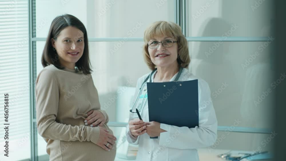 Waist up shot of beautiful pregnant woman and senior female doctor smiling and posing together for camera in clinic Waist up shot of beautiful pregnant woman and senior female doctor smiling and posing together for camera in clinic