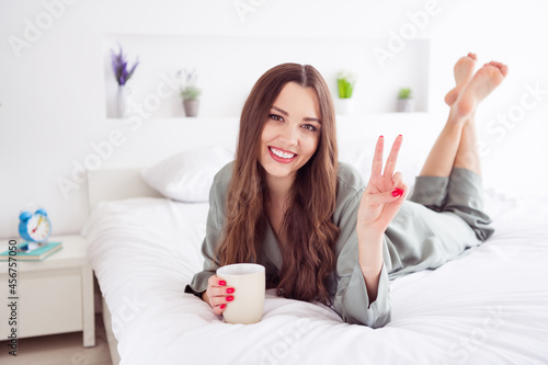 Bild auf Leinwand Portrait of attractive cheerful girl lying on cosy bed drinking cacao showing v-