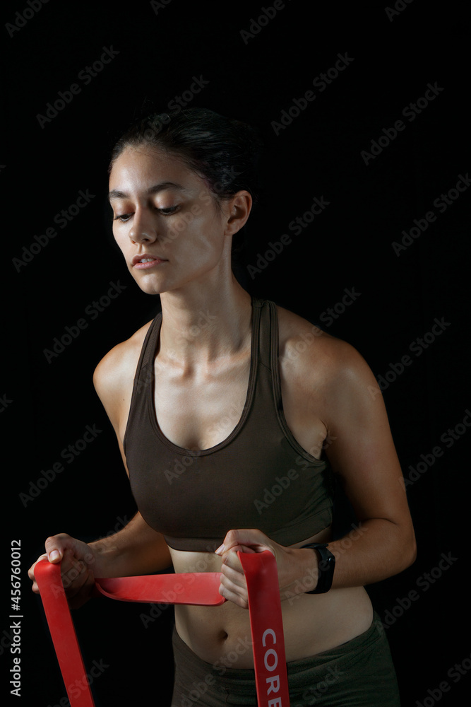 Mujer joven caucásica entrenando bíceps con una banda elástica roja, con  una expresión neutral en la