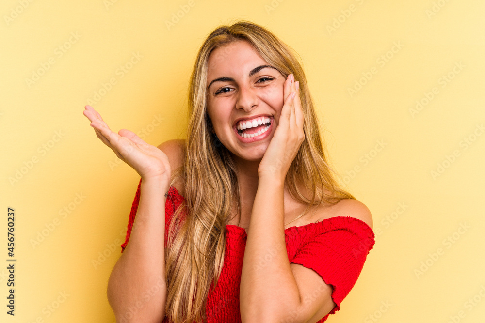 Fototapeta premium Young caucasian blonde woman isolated on yellow background holds copy space on a palm, keep hand over cheek. Amazed and delighted.